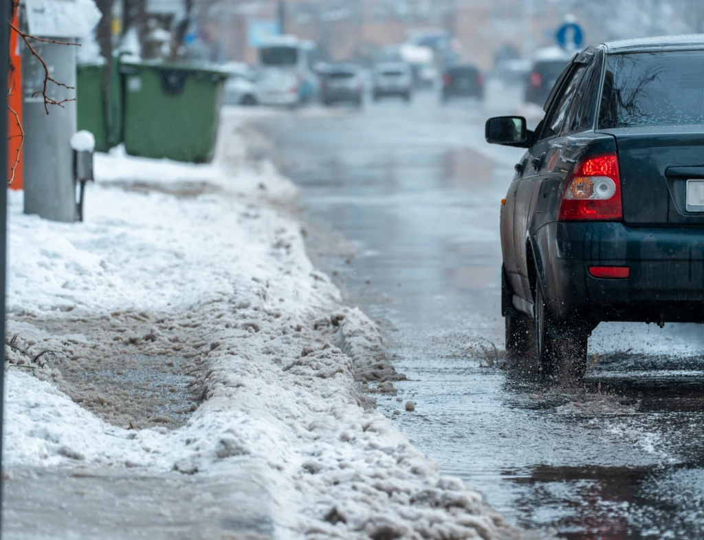 Na początku grudnia temperatura znacząco wzrośnie? Samochód jadący zaśnieżoną i mokrą ulicą w mieście, przy krawężniku widoczne zaspy śniegu, a w tle inne pojazdy i zielone kontenery na odpady.