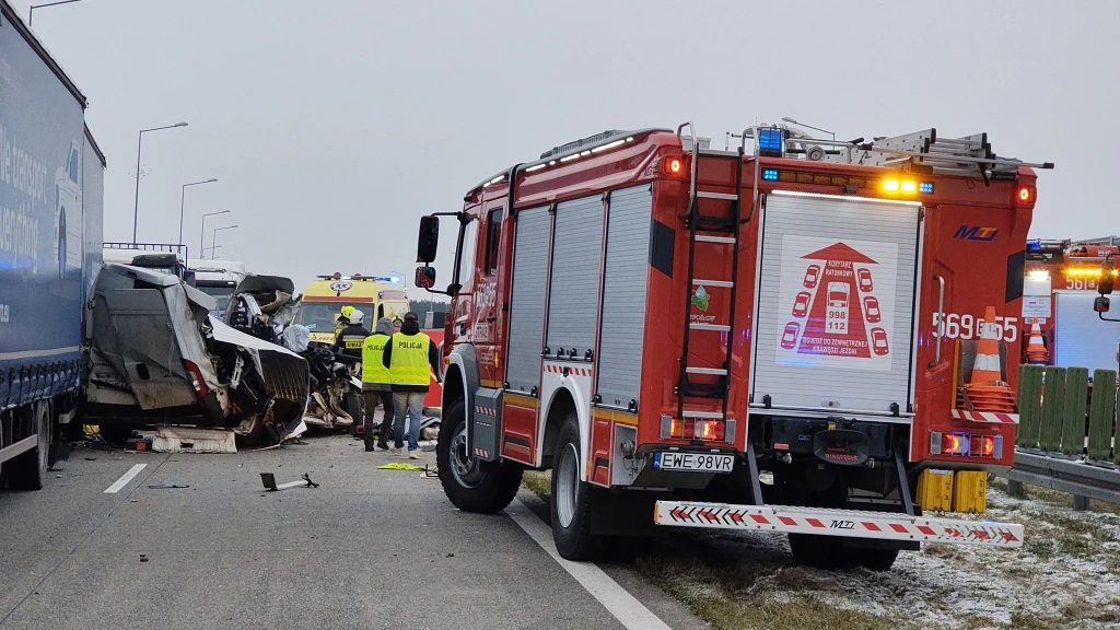 Straż pożarna i ratownicy zabezpieczają miejsce poważnego wypadku drogowego na autostradzie, zniszczony pojazd znajduje się pomiędzy samochodem ciężarowym a samochodem strażackim, wokół panuje zimowa aura.