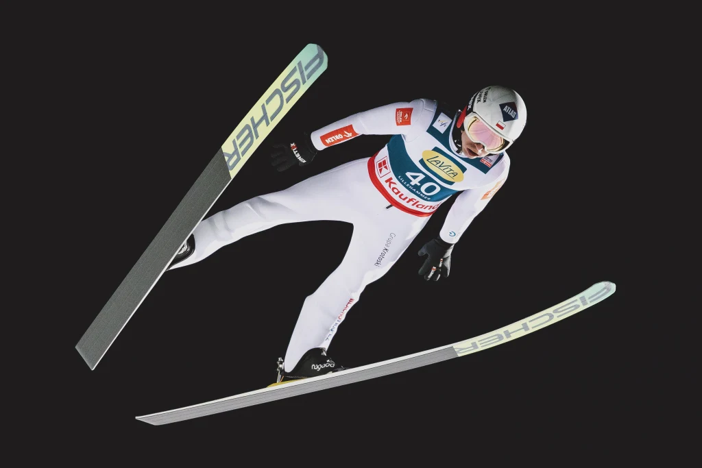 Kamil Stoch podczas Pucharu Świata w skokach narciarskich w Lillehammer Skoczek narciarski ubrany w biały kombinezon i kask znajduje się w powietrzu podczas lotu, z nartami rozłożonymi na boki, na ciemnym tle podkreślającym dynamikę ruchu.