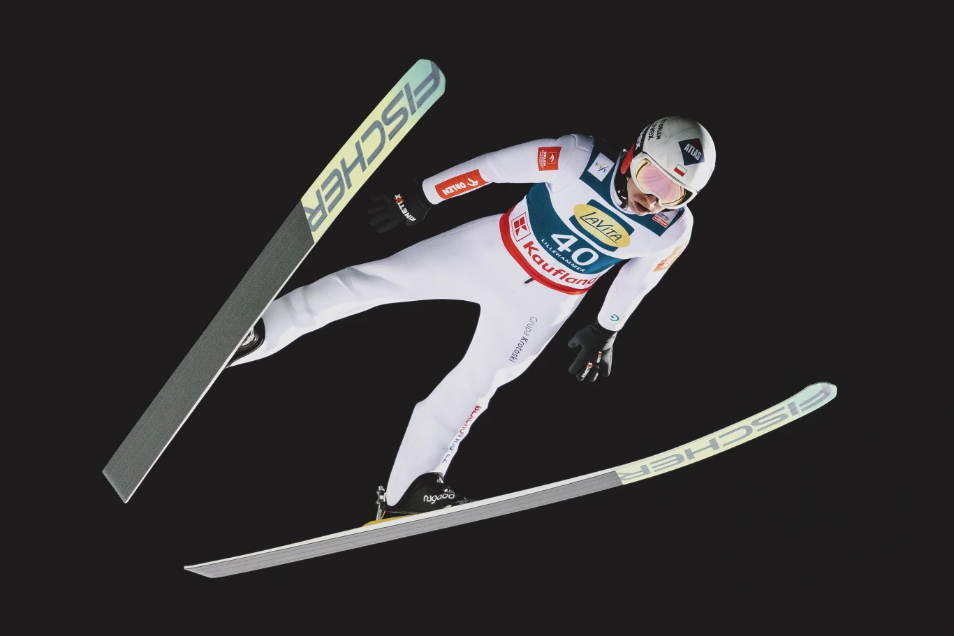 Kamil Stoch podczas Pucharu Świata w skokach narciarskich w Lillehammer Skoczek narciarski ubrany w biały kombinezon i kask znajduje się w powietrzu podczas lotu, z nartami rozłożonymi na boki, na ciemnym tle podkreślającym dynamikę ruchu.