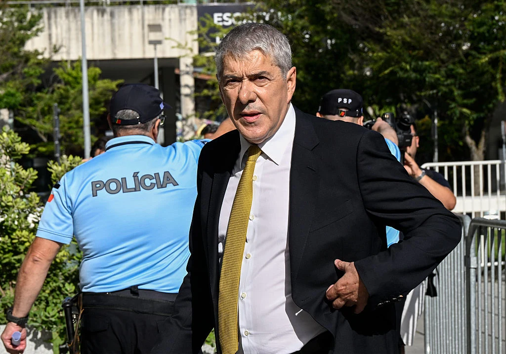 Jose Socrates, były premier Portugalii Mężczyzna w garniturze i żółtym krawacie idzie na tle policjantów ubranych w niebieskie koszule, w otoczeniu zieleni i ogrodzeń.