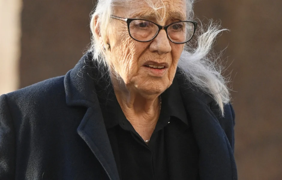 Maja Komorowska Starsza kobieta z siwymi włosami spiętymi do tyłu, ubrana w ciemny płaszcz i okulary, patrzy przed siebie z poważnym wyrazem twarzy. W tle rozmyte, neutralne tło.