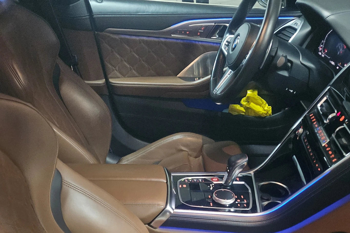 Wnętrze luksusowego samochodu z brązową skórzaną tapicerką, nowoczesną konsolą środkową z licznymi przyciskami i pokrętłem oraz niebieskim podświetleniem. Na fotelu pasażera leży żółta torba.