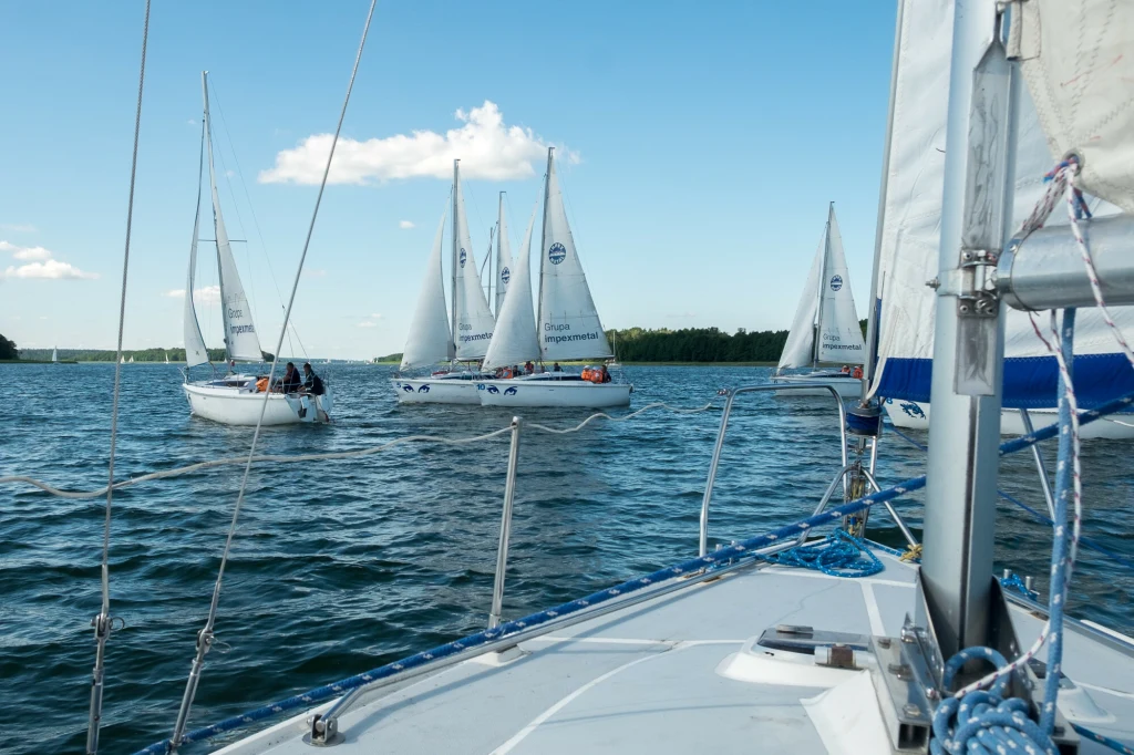 Kilka białych żaglówek płynie po spokojnym jeziorze pod błękitnym niebem z pojedynczymi chmurami, na pierwszym planie widoczny fragment pokładu innej żaglówki, w tle zalesiony brzeg.
