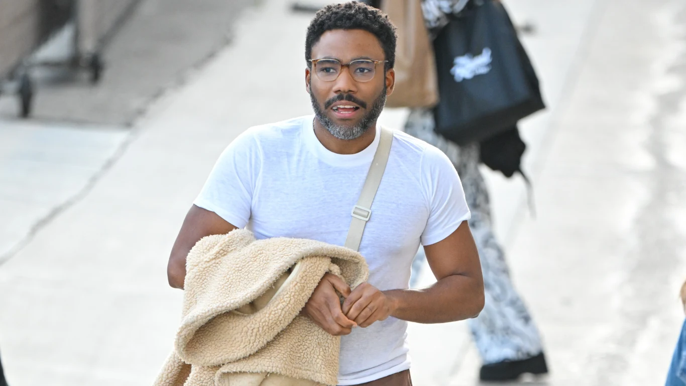 Donald Glover (Childish Gambino) Mężczyzna z okularami i brodą ubrany w białą koszulkę trzyma beżową kurtkę, idzie chodnikiem z torbą przewieszoną przez ramię, w tle widoczne rozmyte postaci i miejski krajobraz.