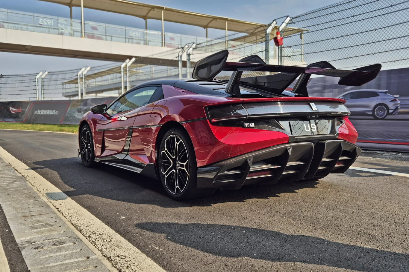 Czerwony samochód sportowy z dużym tylnym spojlerem i czarnymi detalami stoi na torze wyścigowym. W tle widoczne ogrodzenie z siatki oraz fragment trybun. Auto wyróżnia się aerodynamicznym kształtem oraz stylowymi felgami.