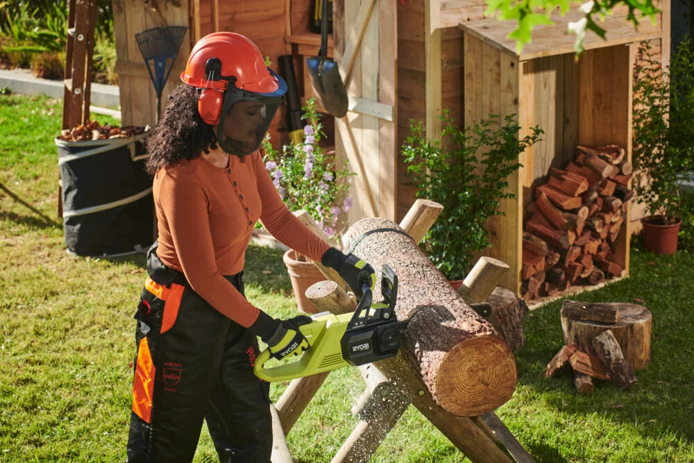 Kobieta w odzieży ochronnej i kasku tnie pilarką łańcuchową duży pień drzewa na stojaku ogrodowym, w tle drewno opałowe, narzędzia ogrodnicze i drewniana szopa.