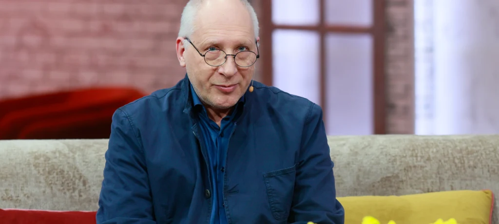 Kuba Sienkiewicz Mężczyzna w okularach i granatowej marynarce siedzi na kanapie ozdobionej kolorowymi poduszkami, w tle rozmyte okno i ceglana ściana.