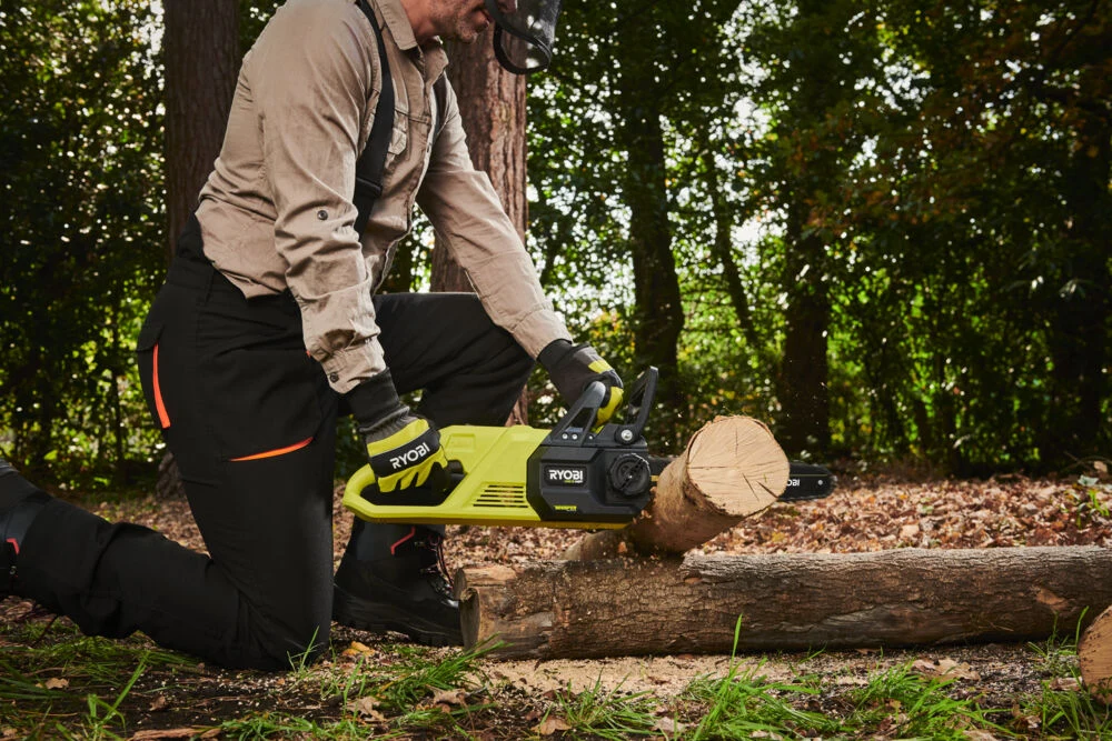 Mężczyzna w ochronnych ubraniach i kasku używa piły łańcuchowej Ryobi do cięcia grubej gałęzi drzewa na tle lasu.