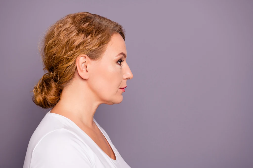 Włosy spięte w kok pasują miedzy innymi do bluz z kapturem Profil kobiety z jasnymi włosami upiętymi z tyłu głowy, ubranej w białą bluzkę na tle szarej jednolitej ściany.