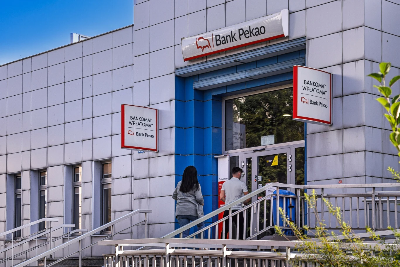 Wejście do oddziału Banku Pekao, dwa banery z logo banku, przed wejściem dwie osoby wchodzące po schodach.