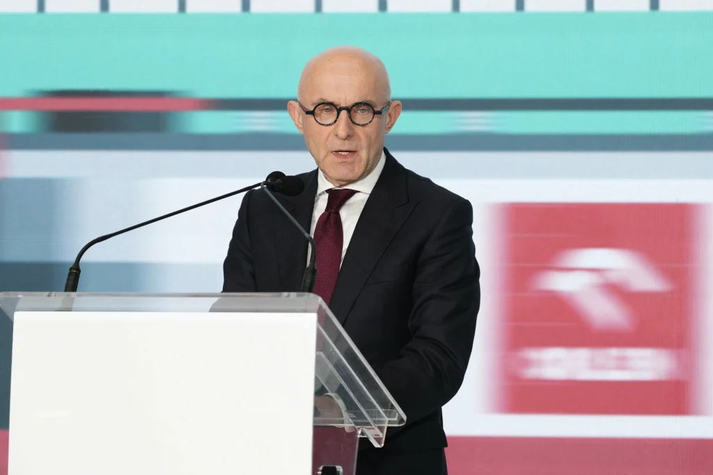 Prezes Orlenu Ireneusz Fąfara Mężczyzna w garniturze i okrągłych okularach przemawia przy przezroczystej mównicy, w tle widoczne są rozmyte logotypy i grafiki na dużym ekranie.