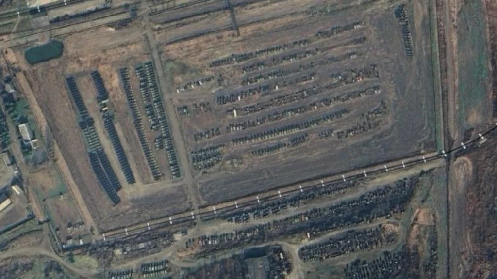 Satelitarne ujęcie dużego placu z licznymi rzędami wojskowych pojazdów i sprzętu ustawionych na otwartym, ogrodzonym terenie. Widać wyraźnie podłużne baraki, tory kolejowe i uporządkowane szeregi sprzętu, tworząc obraz magazynu lub składu wojskowego.