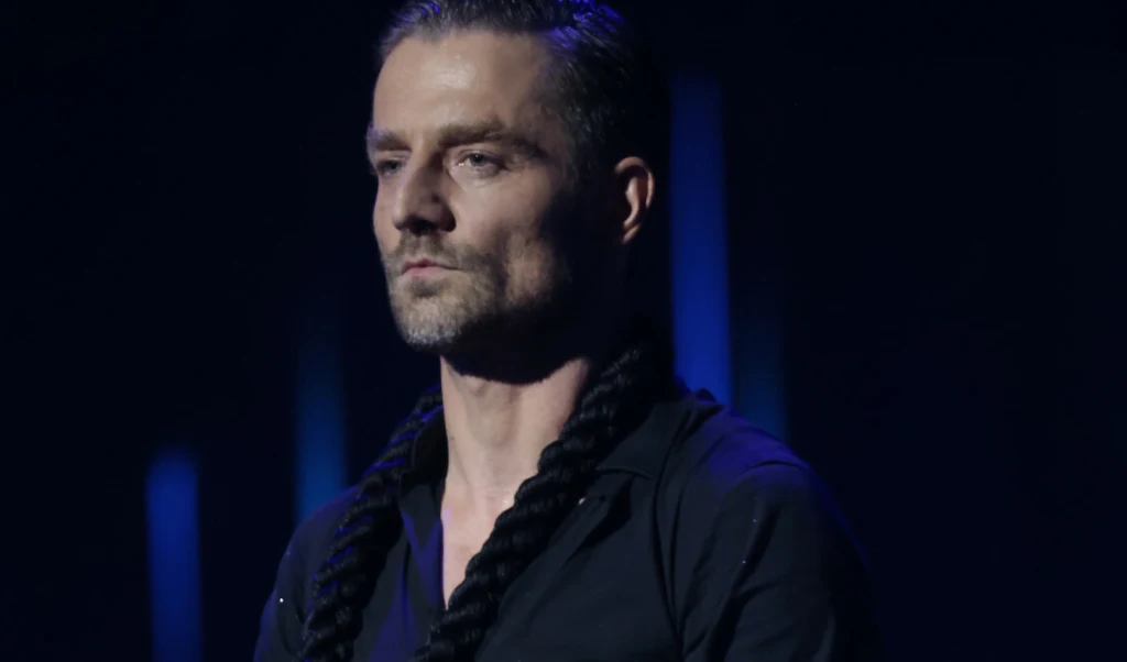 Mężczyzna o poważnym wyrazie twarzy w czarnej koszuli z ciemnym sznurem owiniętym wokół szyi na ciemnym tle.