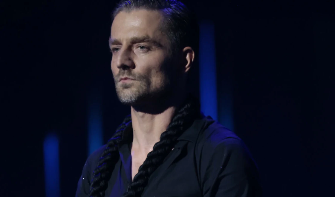 Marcin Rogacewicz Marcin Rogacewicz w ciemnym ubraniu z czarnym sznurem na szyi, na ciemnym tle w niebieskim oświetleniu.
