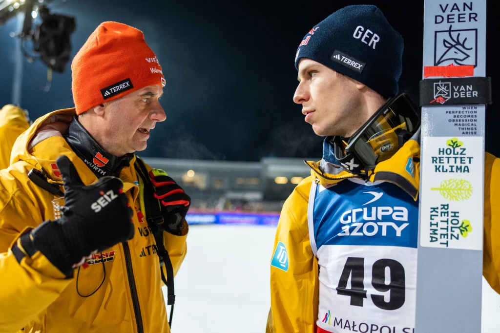Stefan Horngacher i Andreas Wellinger, 21.01.2024 r. Dwóch mężczyzn ubranych w sportowe kurtki prowadzi rozmowę podczas zawodów skoków narciarskich, jeden z nich trzyma narty ze znakiem Van Deer i naklejkami promującymi ochronę klimatu, w tle widoczne trybuny podświetlone sztucznym światłem.
