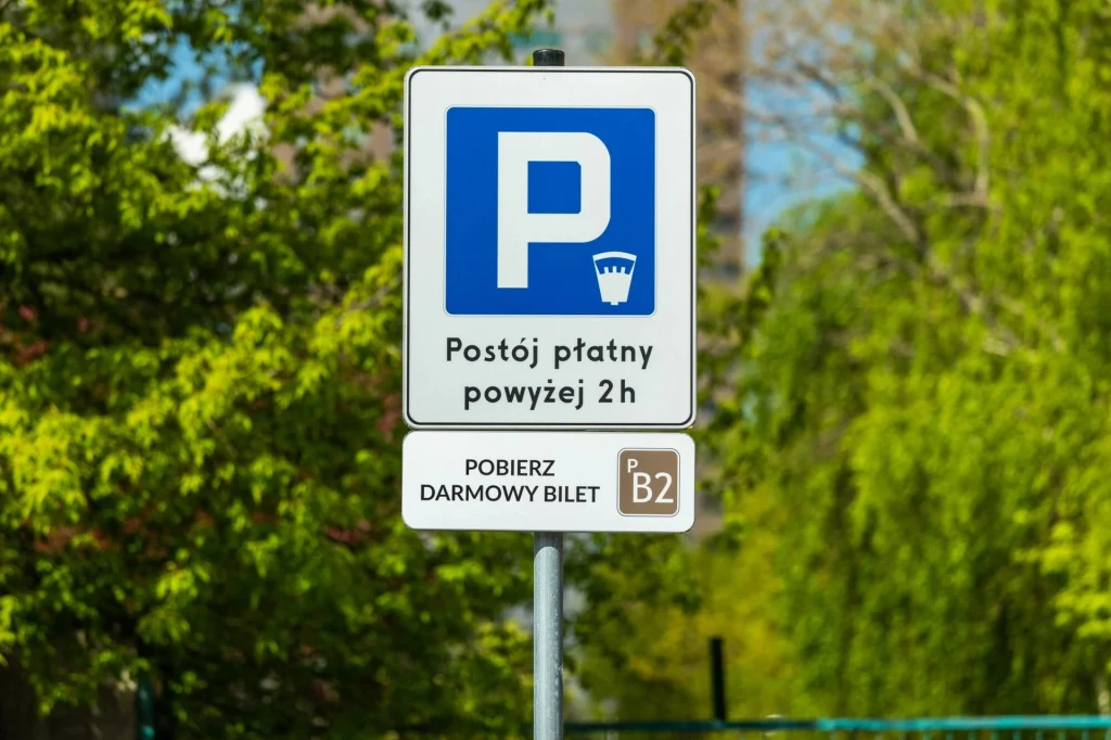 Znak informujący o płatnym parkingu po przekroczeniu dwóch godzin postoju oraz informacja o konieczności pobrania darmowego biletu z oznaczeniem B2, otoczenie zieleni i drzewa w tle.