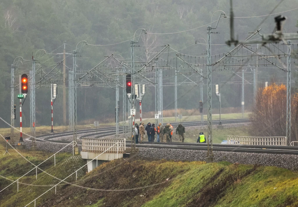 Francuska prasa napisała o aktach dywersji w Polsce. Na zdj. uszkodzone tory w pobliżu stacji PKP Mika Grupa ludzi zgromadzona na torach kolejowych w otoczeniu infrastruktury kolejowej, widoczne sygnalizatory świetlne, przewody trakcyjne oraz elementy techniczne linii kolejowej. Zieleń i drzewa w tle, atmosfera pochmurna.