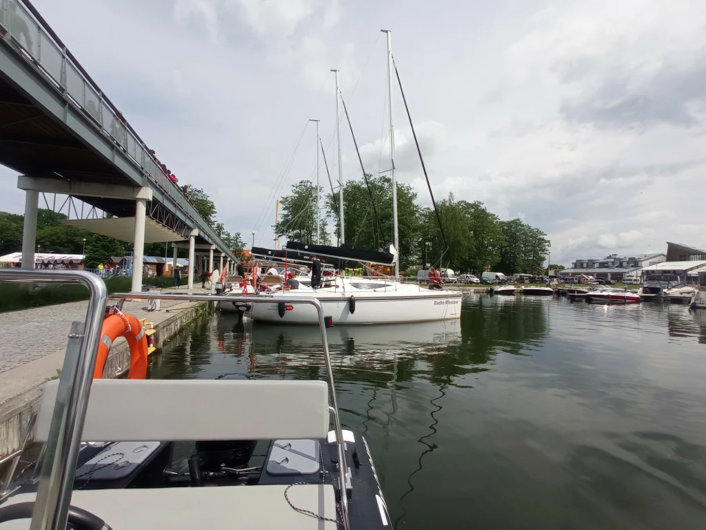 Zacumowane jachty w marinie, widoczne pomosty, łódź motorowa na pierwszym planie oraz zabudowania i zieleń w tle pod pochmurnym niebem.