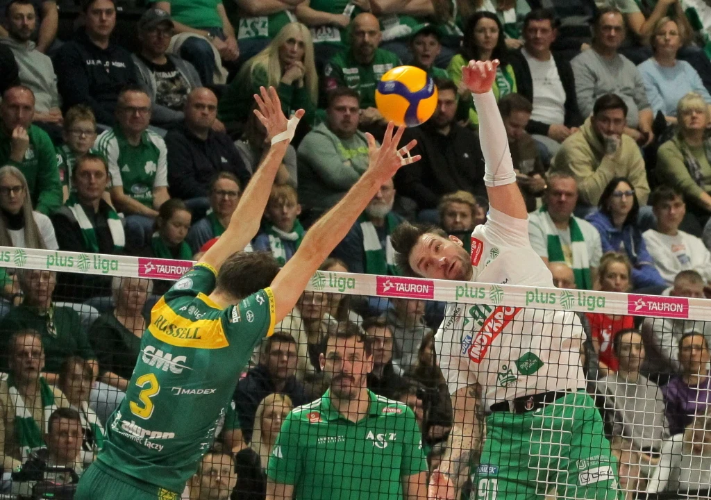 Dwóch siatkarzy podczas dynamicznej akcji przy siatce, jeden z nich próbuje zablokować piłkę po stronie przeciwnika, wokół widoczna licznie zgromadzona publiczność w hali sportowej.