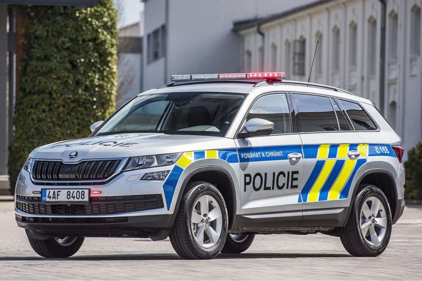 Z zewnątrz standardowy policyjny Kodiaq, ale zaskoczenie kryje się pod maską. Radiowóz czeskiej policji marki Škoda Kodiaq zaparkowany na brukowanej ulicy, otoczony budynkami o jasnych elewacjach i zielenią w tle, widoczne oznaczenia Policie oraz sygnalizator świetlny na dachu.