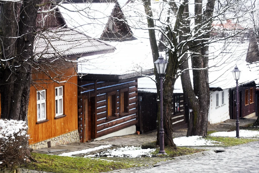 Drewniane chaty pokryte śniegiem, ustawione wzdłuż brukowanej uliczki, otoczone nagimi drzewami oraz klasycznymi latarniami, dające wrażenie spokojnej, zimowej scenerii w małej miejscowości.