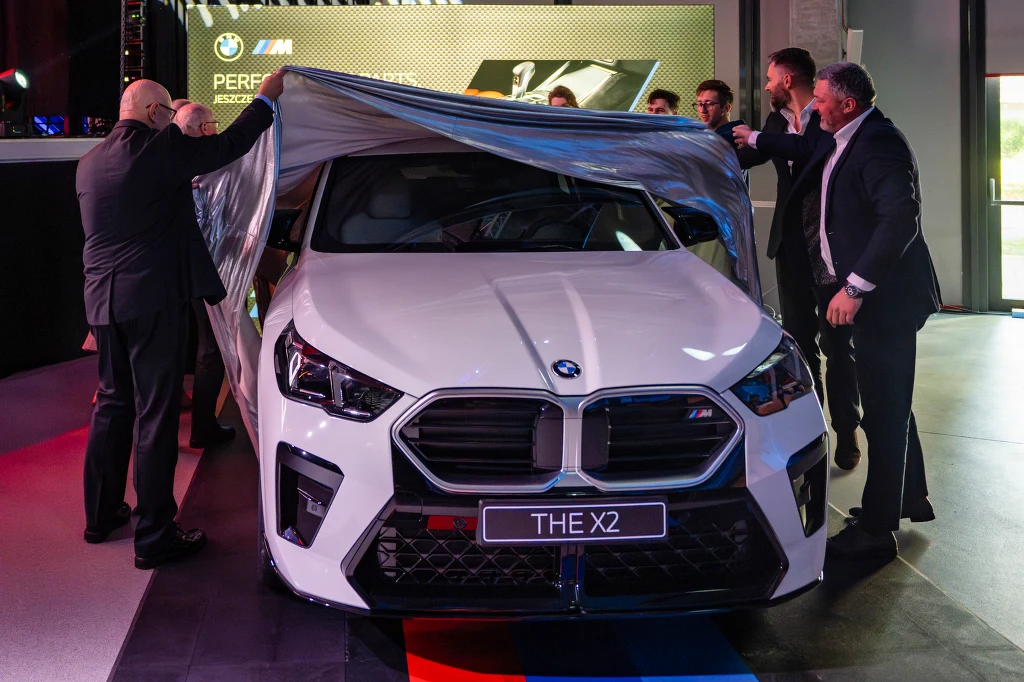 Biały samochód BMW X2 odsłaniany przez kilku elegancko ubranych mężczyzn podczas oficjalnej prezentacji, w tle widoczna scena oraz logo marki.
