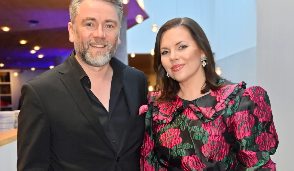 Kuba Badach, i Aleksandra Kwaśniewska Para ubrana elegancko, mężczyzna w czarnym garniturze oraz kobieta w ciemnej, wzorzystej sukience z motywem róż, stoją blisko siebie w jasnym, nowoczesnym wnętrzu. Oboje uśmiechnięci, przyjazna atmosfera.