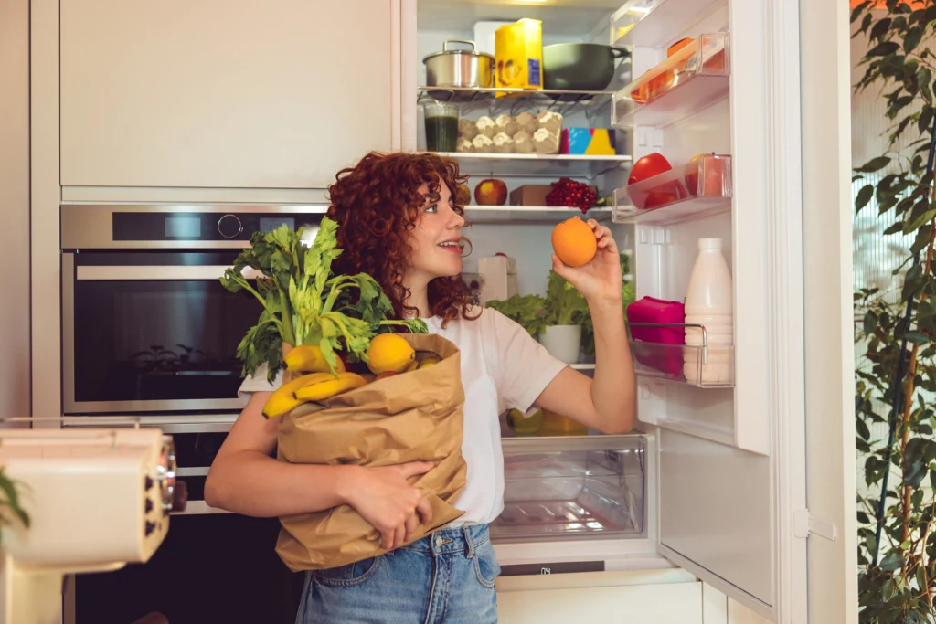 Dobrze przyjrzyj się temu, jak przechowujesz warzywa i owoce. To właśnie tu tkwi sekret ich długiej świeżości Kobieta trzyma torbę z warzywami i owocami, wyciąga pomarańczę stojąc przed otwartą lodówką w nowoczesnej kuchni. W lodówce widoczne są świeże produkty spożywcze.