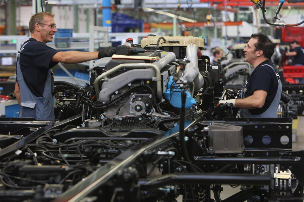 Dwóch mężczyzn pracuje przy montażu dużych silników na hali produkcyjnej, otoczeni elementami maszyn oraz narzędziami, atmosfera współpracy i skupienia.