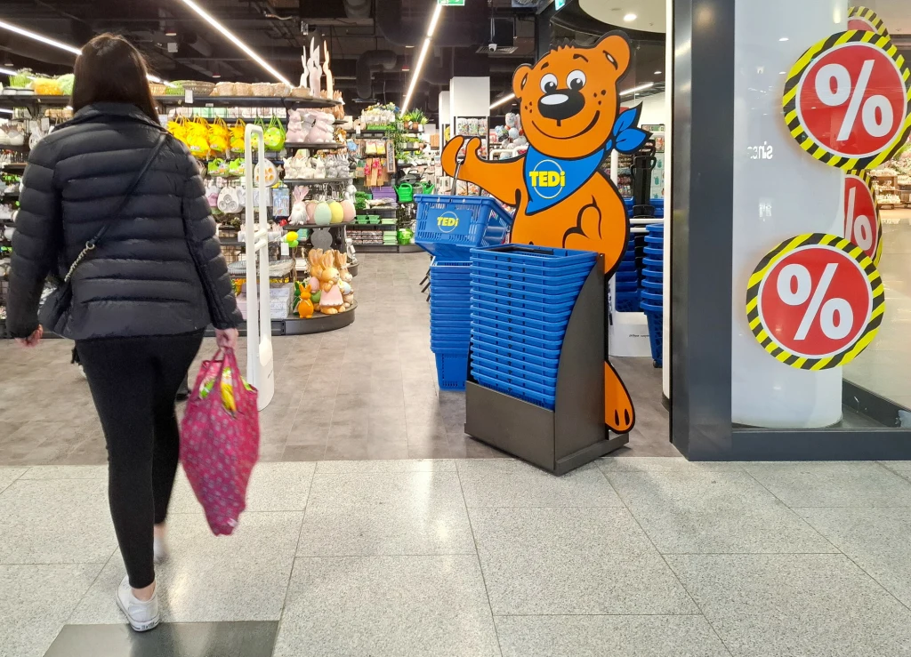 Sieć Tedi wydała pilny komunikat ws. produktów wycofanych ze sprzedaży Kobieta z różową torbą na ramieniu wchodzi do sklepu, przy wejściu widoczny niebieski stojak z koszykami z kartonową maskotką misia TEDi i półki z różnorodnym asortymentem. Po prawej stronie przy wejściu znajdują się oznaczenia promocji procentowych.