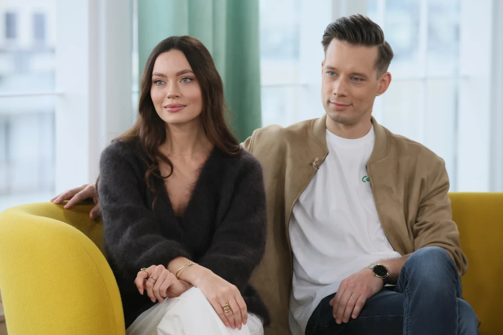 Dwoje młodych ludzi siedzi na żółtej sofie, kobieta z długimi ciemnymi włosami w ciemnym swetrze i mężczyzna w beżowej kurtce oraz białej koszulce patrzą w jednym kierunku, w tle jasne okno i zielona zasłona.