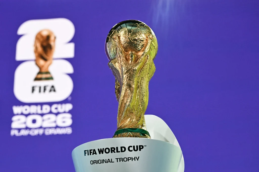 Złota statuetka Pucharu Świata FIFA na białym podeście z napisem „FIFA World Cup Original Trophy” na tle fioletowej tablicy promującej Mistrzostwa Świata FIFA 2026.
