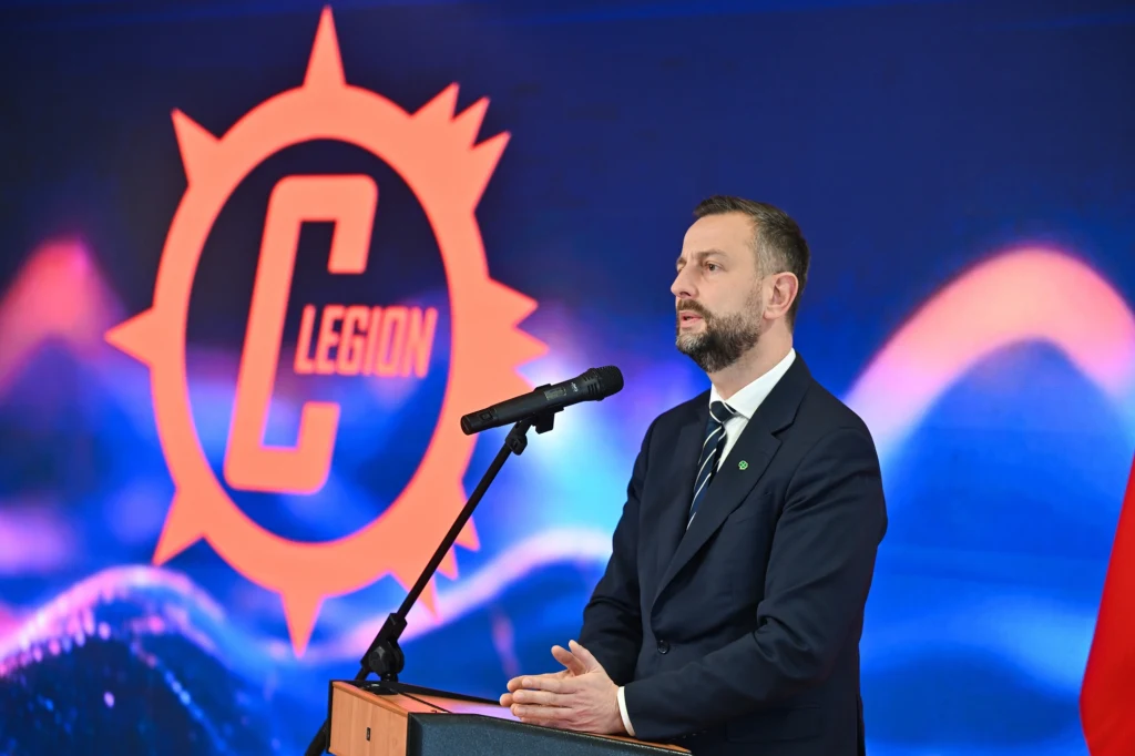 Mężczyzna w garniturze przemawia przy mównicy na tle podświetlonego logotypu z napisem 'C Legion'.