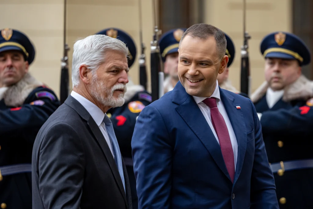 Dwóch mężczyzn w garniturach rozmawia ze sobą na tle żołnierzy w mundurach galowych stojących w szeregu, jeden z nich uśmiecha się, panuje oficjalna atmosfera.