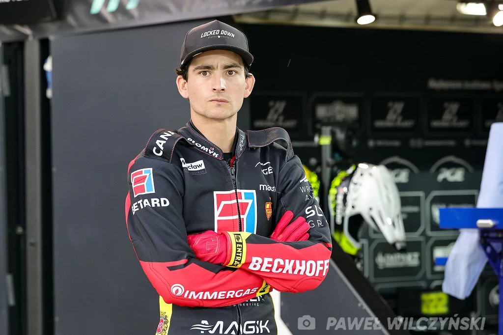 Młody żużlowiec w pełnym stroju sportowym i czapce stoi z założonymi rękami na tle parku maszyn, za nim widać sprzęt sportowy oraz tablice z logotypami sponsorów.