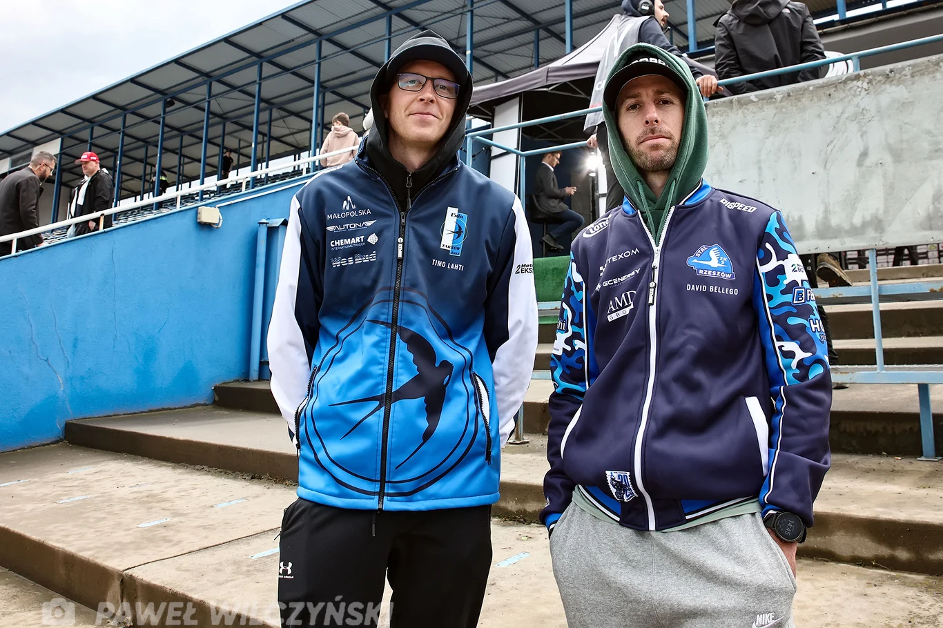 Timo Lahti i David Bellego. Dwóch młodych mężczyzn w sportowych bluzach i kapturach stoi obok siebie na betonowych trybunach stadionu, wokół nieliczni widzowie. W tle widać zadaszenie trybun oraz niebieskie barierki.