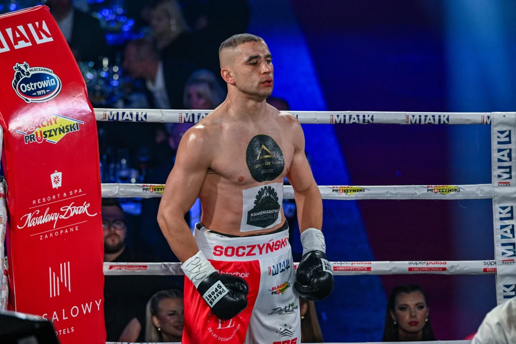 Bokser stojący w ringu z zaciśniętymi pięściami, na jego ciele widoczne tatuaże i reklamy sponsorów. Otaczają go liny ringu oraz reklamy promujące wydarzenie, w tle obecni są widzowie i sędziowie.