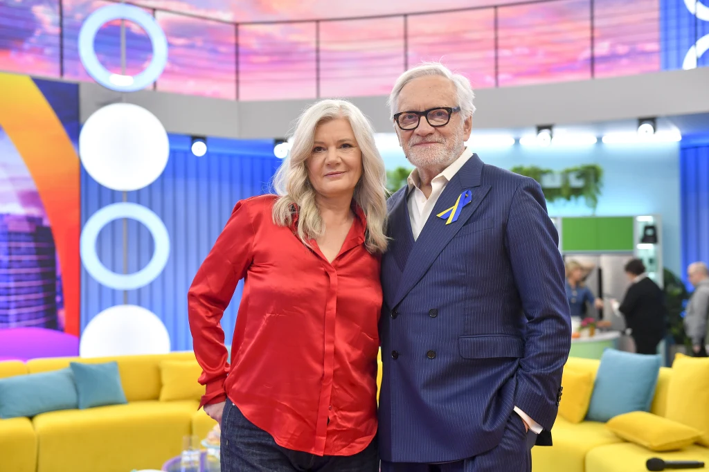 Para eleganckich, starszych osób pozuje razem w kolorowym, nowoczesnym wnętrzu. Kobieta ubrana w czerwoną, satynową bluzkę, mężczyzna w niebieskim garniturze z przypiętą wstążką w barwach niebiesko-żółtych, co może nawiązywać do wsparcia dla Ukrainy. Z...