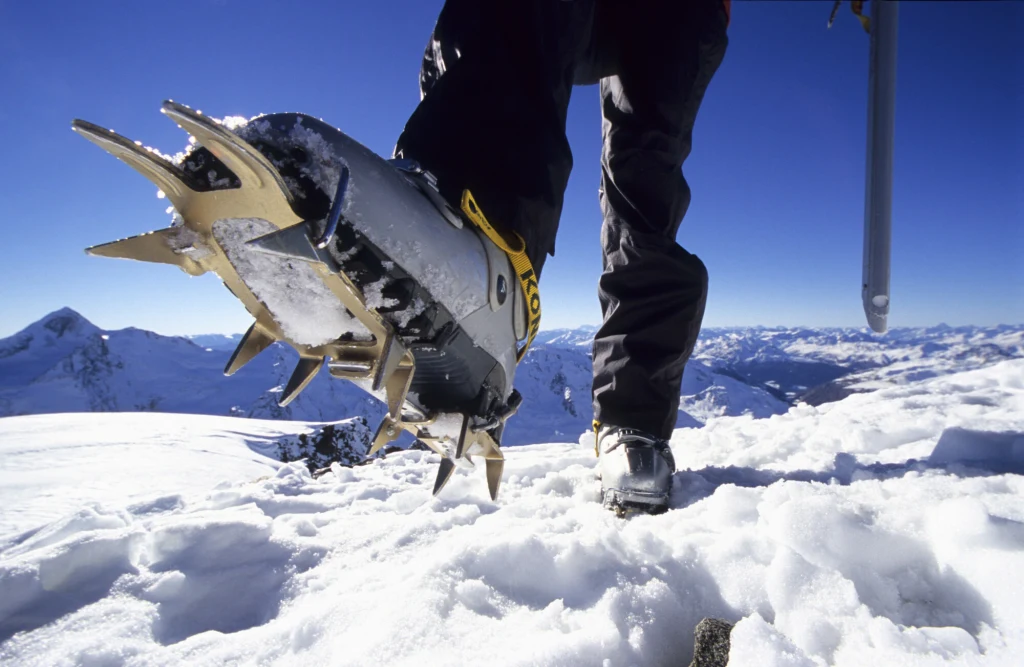 Buty wyposażone w raki do wspinaczki na śnieżnej powierzchni gór, wyeksponowany detal ostrzy, w tle rozległy krajobraz ośnieżonych gór i błękitne niebo.