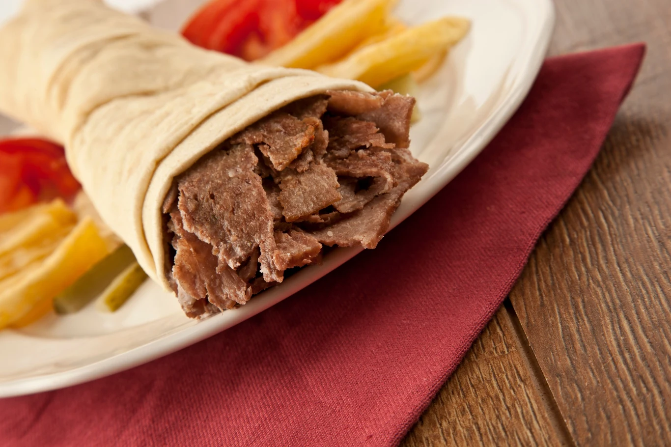 Kebab w placku lavash z kawałkami mięsa, podany na białym talerzu. W tle widoczne plastry pomidora i frytki. Całość znajduje się na czerwonej serwetce ułożonej na drewnianym stole.
