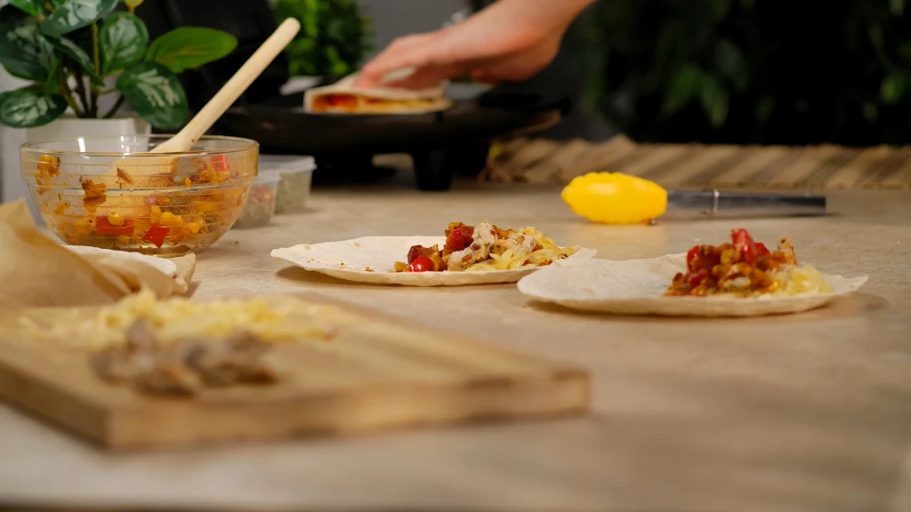 Na stole leżą dwie tortille z farszem warzywnym oraz serem, w tle miska ze świeżo przygotowanym nadzieniem, roślina ozdobna i ręka osoby układającej kolejną tortillę.