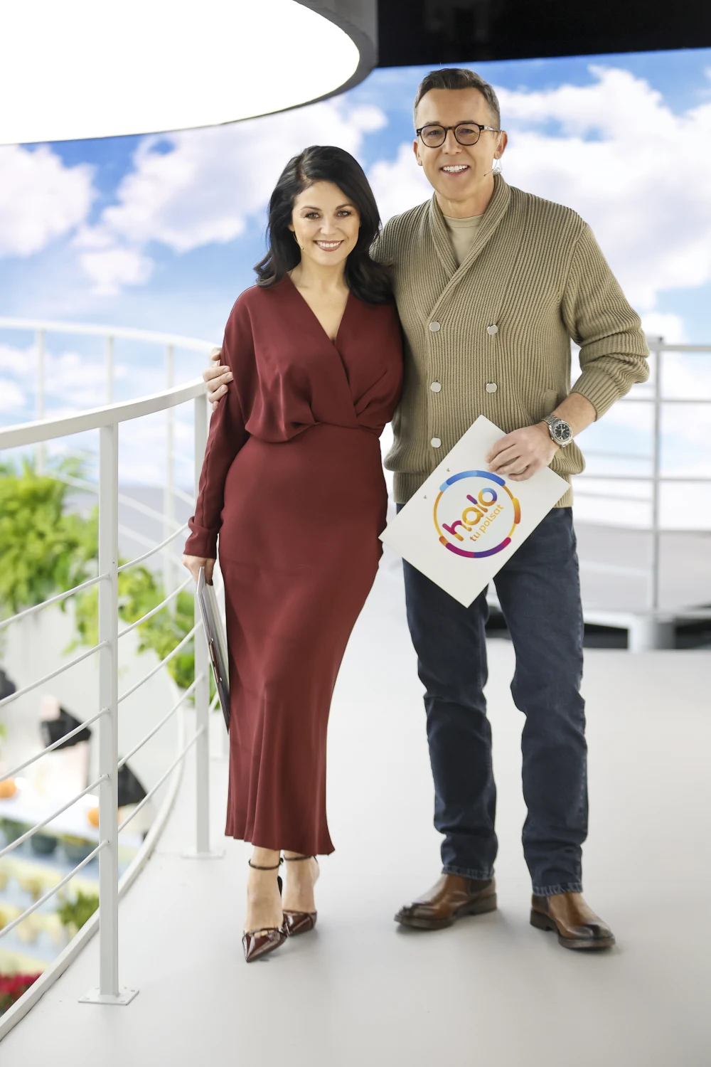 Dwie elegancko ubrane osoby stojące obok siebie na tle jasnego wnętrza z elementami roślinności i panoramicznego widoku na niebo, trzymające w rękach materiały programowe.