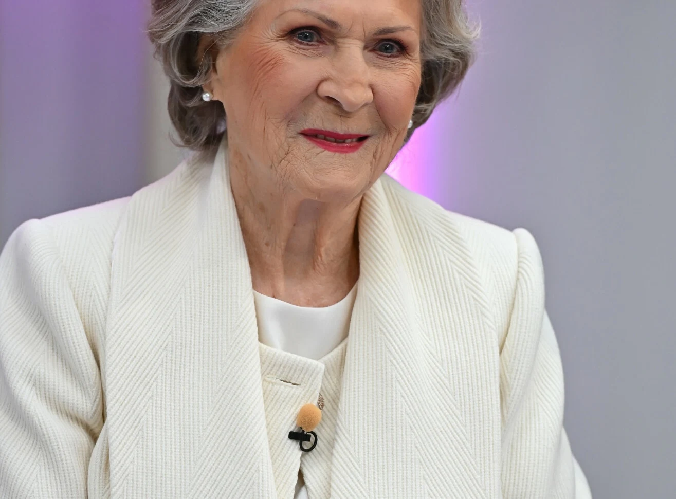 Powstałą aleja imienia Ireny Santor Starsza kobieta z krótkimi, siwymi włosami w eleganckiej, jasnej marynarce, uśmiechnięta, z delikatnym makijażem, siedzi w dobrze oświetlonym wnętrzu z fioletowym podświetleniem w tle.