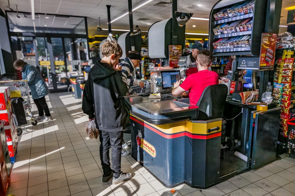 Scena w supermarkecie, gdzie kasjer w czerwonej koszulce obsługuje młodego chłopca stojącego przy ladzie, po bokach półki pełne produktów, w tle widoczni inni klienci z wózkami zakupowymi.