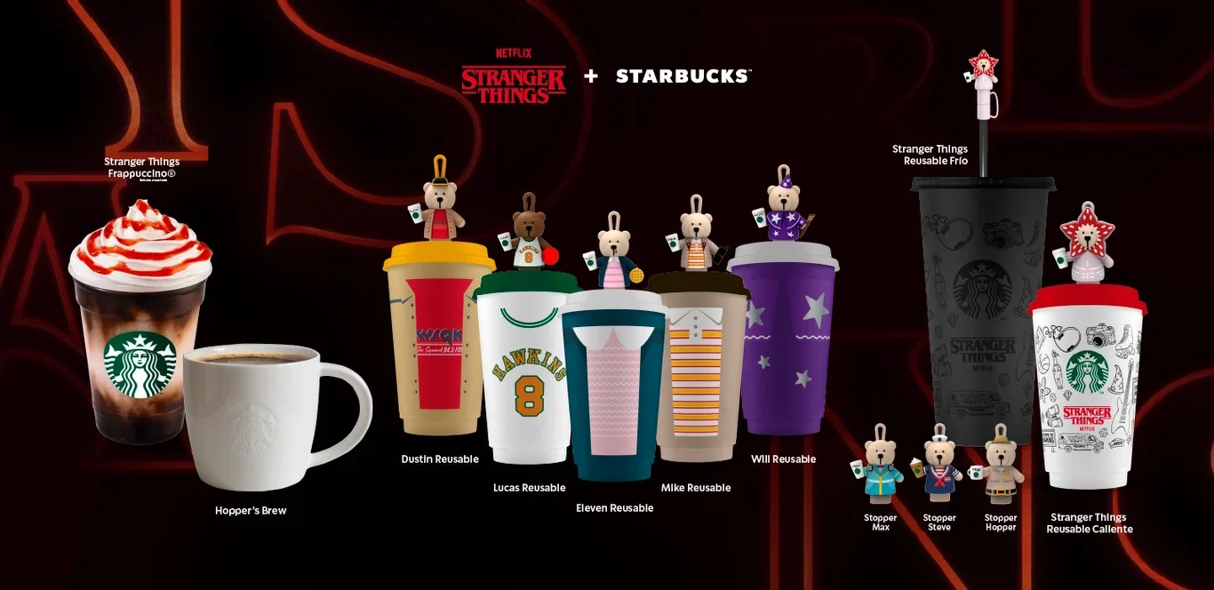 Rząd kolorowych kubków Starbucks z motywami serialu Stranger Things, ozdobionych figurkami przedstawiającymi postacie z serialu, dwie filiżanki z różnymi napojami po lewej stronie oraz czarne i białe kubki z dodatkowymi ozdobami po prawej stronie, cało...