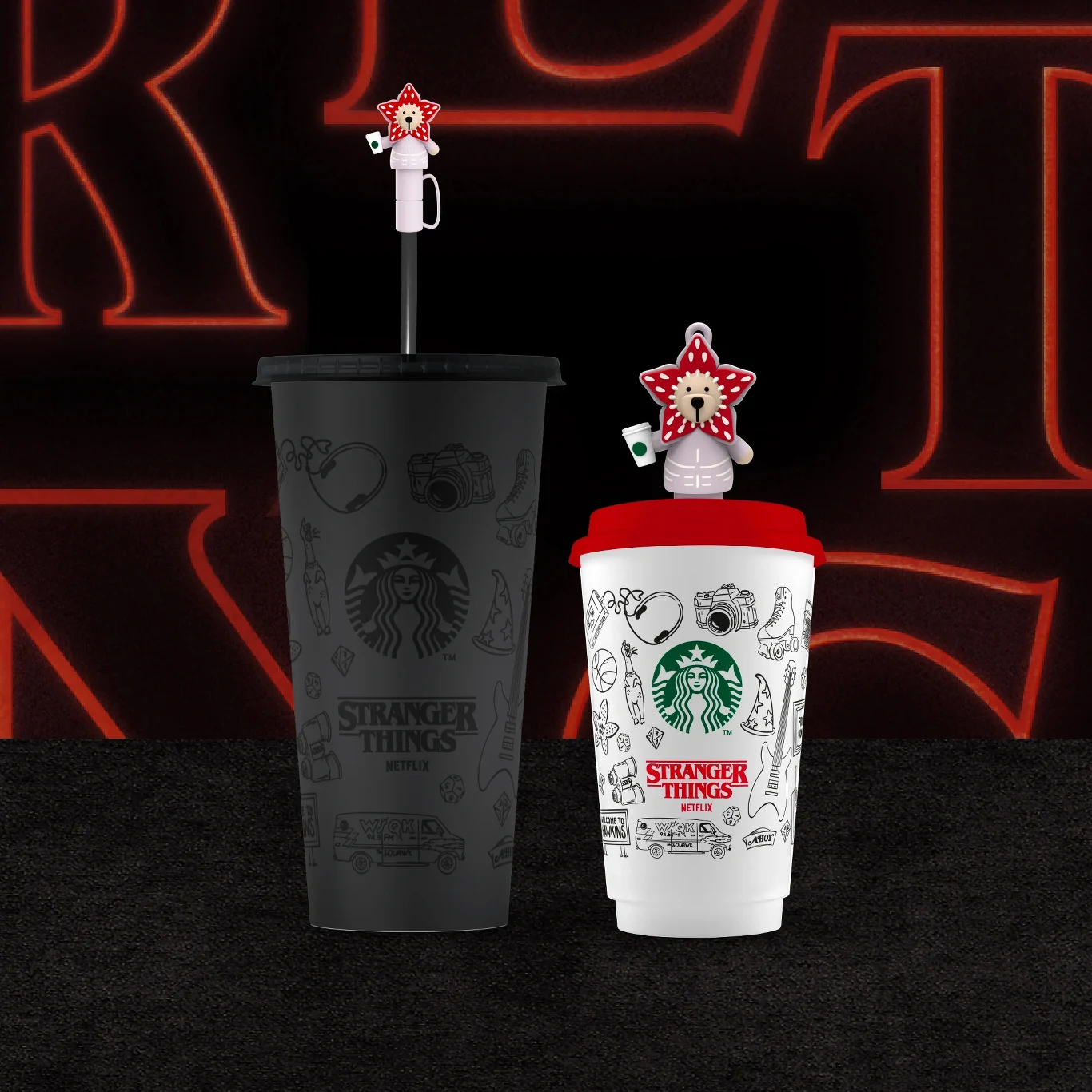 Dwa kubki Starbucks z edycji Stranger Things, jeden czarny z czarną słomką i figurką potwora Demogorgon, drugi biały z czerwoną pokrywką i słomką z identyczną figurką, oba ozdobione tematycznymi grafikami związanymi z serialem.