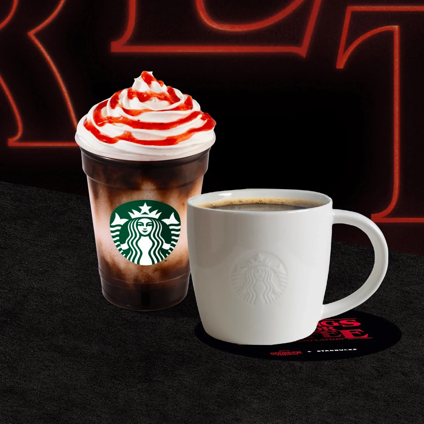 Plastikowy kubek z logo Starbucks wypełniony kawą z bitą śmietaną i polewą truskawkową obok białej filiżanki z kawą ustawionej na czarnej podstawce. W tle czerwone litery, które są rozmyte.