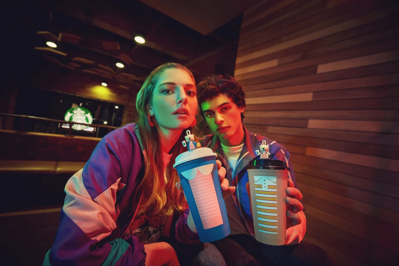 Dwie młode osoby w sportowych kurtkach siedzą w lokalu o nowoczesnym wystroju i trzymają w dłoniach ozdobione kubki napojów. Oświetlenie jest kolorowe i tworzy dynamiczny, miejski klimat. Za nimi widoczny jest fragment logo znanej kawiarni.
