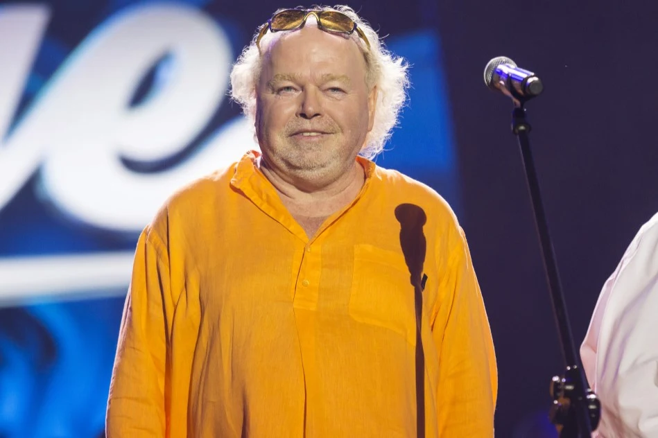 Stanisław Wenglorz był gwiazdą w PRL-u Mężczyzna z siwymi włosami i okularami przeciwsłonecznymi na głowie ubrany w pomarańczową koszulę stoi na scenie obok mikrofonu. Tło rozmyte, utrzymane w ciemnych kolorach.